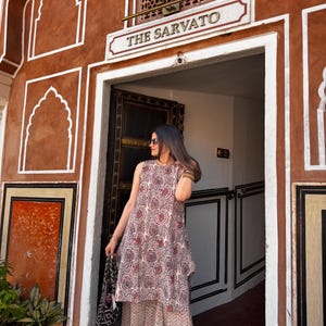 May include: A sleeveless, patterned dress with a matching skirt. The dress features a red and white paisley design. The person is holding a black and white tote bag. The background is a brown building with the text "THE SARVATO".
