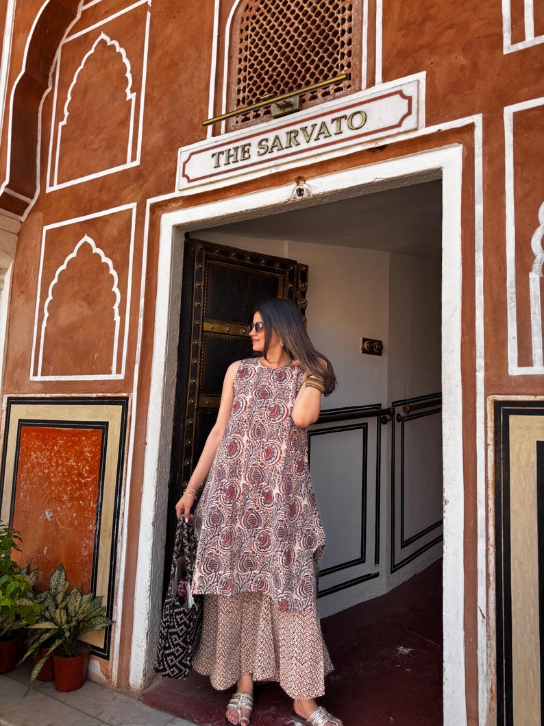 May include: A sleeveless, patterned dress with a matching skirt. The dress features a red and white paisley design. The person is holding a black and white tote bag. The background is a brown building with the text "THE SARVATO".