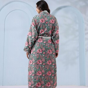 May include: A long, floral-print robe in shades of teal and pink. The robe features a repeating pattern of large pink flowers and green leaves. It has long sleeves, a white belt, and is worn over white palazzo pants.