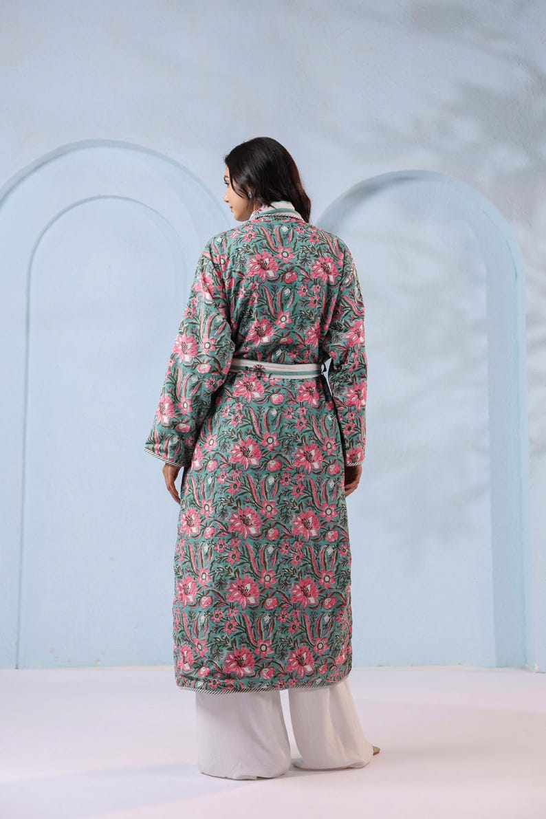 May include: A long, floral-print robe in shades of teal and pink. The robe features a repeating pattern of large pink flowers and green leaves. It has long sleeves, a white belt, and is worn over white palazzo pants.