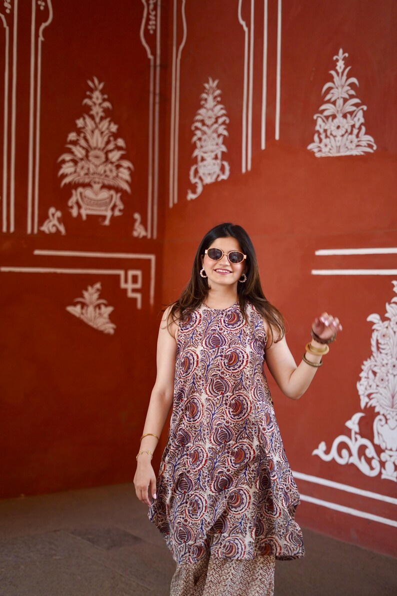 May include: A sleeveless dress and matching pants with a paisley pattern in shades of red, blue, and beige. The outfit is worn in a room with red walls and white decorative accents. The person is wearing sunglasses and bracelets.