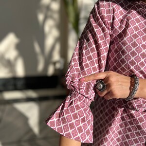 May include: A pink and white patterned top with a ruffled sleeve. The top has a geometric design with a silver ring on the finger of the hand wearing the top.
