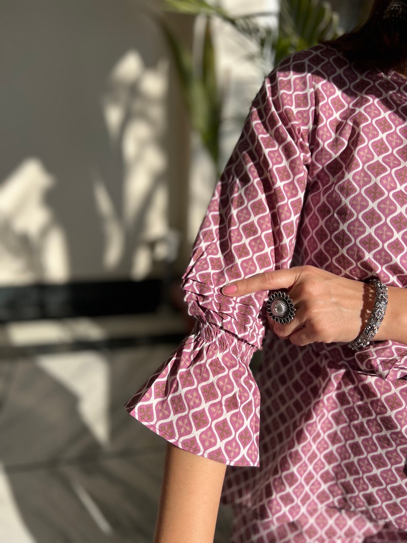 May include: A pink and white patterned top with a ruffled sleeve. The top has a geometric design with a silver ring on the finger of the hand wearing the top.