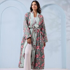 May include: A long, floral-print robe in shades of green and pink, with a striped accent and a matching belt. The robe is worn over white pants and a white top. The outfit is completed with gold sandals.