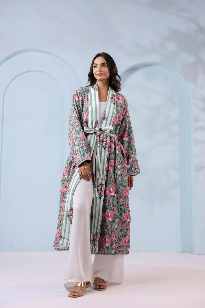 May include: A long, floral-print robe in shades of green and pink, with a striped accent and a matching belt. The robe is worn over white pants and a white top. The outfit is completed with gold sandals.