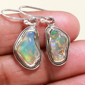 May include: A pair of silver earrings with iridescent opal stones. The opals have a unique, irregular shape and are set in a silver bezel. The earrings are suspended from silver ear wires. The text "SilverHubJewels" is in the top left corner.