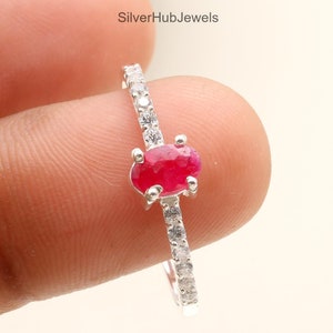 Op de afbeelding: Een zilveren ring met een helderrode ovale edelsteen in het midden, geflankeerd door kleinere, heldere stenen. De ringband is versierd met extra heldere stenen. De ring wordt tegen een lichte achtergrond gehouden.