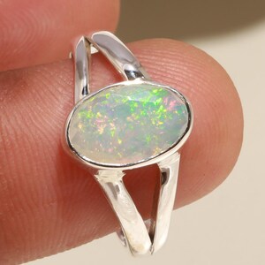 May include: A silver ring featuring an oval opal gemstone. The opal displays a play of color with iridescent green and pink hues. The ring's band has a split design, creating an open, modern look. The ring is held by a person.