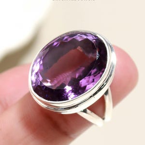 May include: A silver ring featuring a large, oval-shaped amethyst gemstone. The amethyst is a rich purple hue, set in a simple silver band. The ring is held by a finger. The text "SilverHubJewels" is visible in the upper left corner.