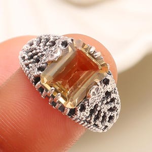 May include: A silver ring featuring an amber-coloured rectangular gemstone. The ring has a detailed, textured design around the gemstone and along the band, with small black accents. The ring is held by a finger.
