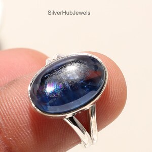 Op de afbeelding: Een zilveren ring met een ovale, donkerblauwe edelsteen. De edelsteen heeft iriserende kwaliteiten en reflecteert meerdere kleuren. De band van de ring is aan de bovenkant gesplitst, wat een uniek ontwerp creëert. De tekst "SilverHubJewels" is zichtbaar.