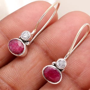Pode incluir: Um par de brincos de prata com uma pedra preciosa ovalada de cor rubi e uma pequena pedra preciosa redonda e transparente. Os brincos têm um design simples com fecho de gancho.