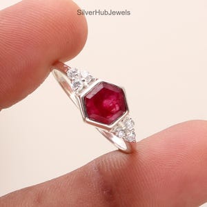Pode incluir: Um anel de prata com uma pedra preciosa hexagonal, vermelho escuro, como peça central, ladeada por pedras menores e transparentes. O anel tem uma banda fina, com o texto "SilverHubJewels" visível.