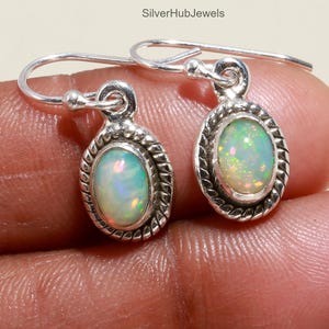 May include: A pair of silver-toned oval earrings with iridescent opal stones. The opals have a mix of green, yellow, and white hues. The earrings have a rope-like border around the opal and a simple hook closure.