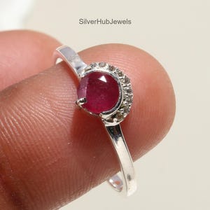 May include: A silver ring featuring a vibrant red gemstone, accented by a halo of smaller, clear stones. The ring's band is a simple, polished silver. The text "SilverHubJewels" is visible.