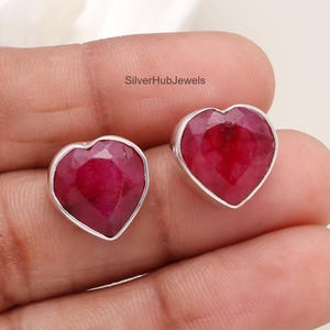 Pode incluir: Um par de brincos de rubi em forma de coração. Cada brinco apresenta uma pedra preciosa vermelha escura facetada em uma moldura prateada. O texto "SilverHubJewels" é visível.
