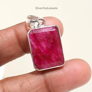 Puede incluir: Un colgante rectangular con una gema rosa intenso y vibrante engastada en un marco plateado. La gema tiene una superficie facetada que capta la luz. El colgante tiene un pequeño lazo en la parte superior para una cadena. El texto "SilverHubJewels" es visible.