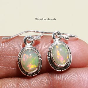 May include: A pair of silver earrings with oval-shaped opal gemstones. The opals display a mix of colors, including green, yellow, and pink. The earrings have a decorative border around the gemstones and a hook closure. The text "SilverHubJewels" is visible.