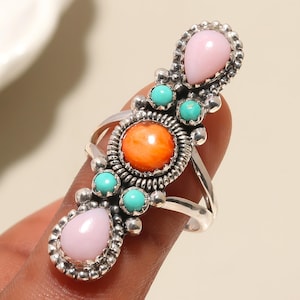 Puede incluir: Un anillo plateado con un diseño alargado, con una piedra central naranja, turquesas y piedras rosas. El anillo tiene un engaste ornamentado y detallado, que muestra un estilo del suroeste o nativo americano.