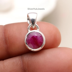 Puede incluir: Un colgante de plata con una piedra preciosa de rubí rojo oscuro y redondo. El rubí está engastado en un bisel y el colgante tiene un diseño sencillo y elegante. El texto "SilverHubJewels" es visible en la imagen.