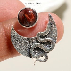 Puede incluir: Un pendiente de plata con un diseño de media luna con superficie texturizada y una serpiente enroscada. Una gema redonda de color marrón rojizo se encuentra sobre la luna. El pendiente está sostenido por un dedo.