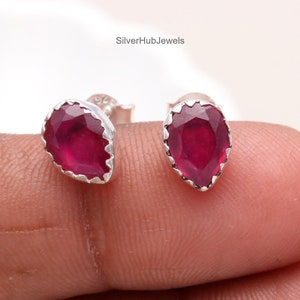 May include: A pair of stud earrings featuring faceted, pear-shaped deep red rubies set in scalloped silver bezels. The earrings are marked with the text SilverHubJewels in the background. Each ruby displays a vibrant, translucent crimson hue.