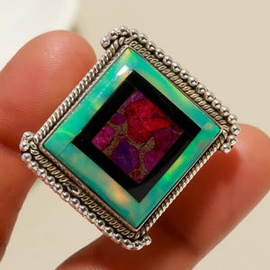 May include: A silver ring with a large, square gemstone. The gemstone features a central black square with a mosaic of red and purple, surrounded by a green and iridescent border. The ring has a beaded edge.