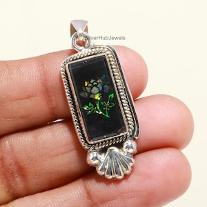 May include: A rectangular silver pendant with a black inlay featuring a floral design. The pendant has a decorative rope-like border and a shell-shaped bottom. The floral design includes green and iridescent accents.