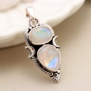 Puede incluir: Colgante de plata con dos cabujones de piedra lunar iridiscente. La piedra superior es ovalada y la inferior en forma de lágrima. El colgante tiene un borde decorativo en forma de cuerda y detalles de media luna.
