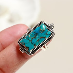 May include: A rectangular silver ring featuring a vibrant turquoise stone with copper-colored matrix patterns. The stone is set within a detailed, textured silver frame. The ring is displayed on a finger.