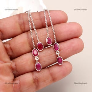 May include: A pair of silver dangle earrings with three oval-shaped ruby gemstones. The earrings have a delicate chain design.