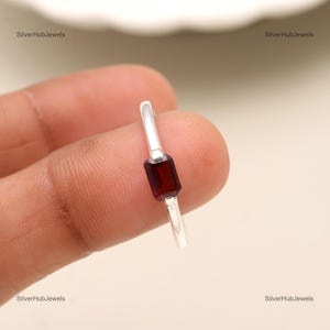 May include: A silver ring with a rectangular red gemstone. The ring is on a finger.