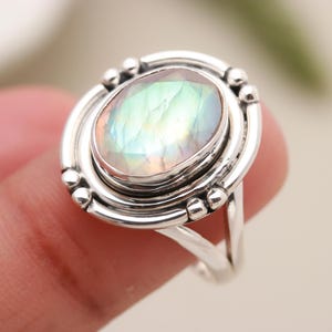 Puede incluir: Anillo de plata con una piedra preciosa ovalada e iridiscente. La piedra preciosa está engastada en un marco circular detallado con pequeños detalles de cuentas. La banda del anillo se divide en dos en la base, creando un diseño único.