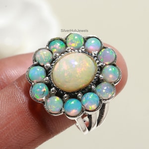May include: A silver ring with a large white opal surrounded by smaller opals in shades of blue and green. The ring is set in a flower-shaped design.