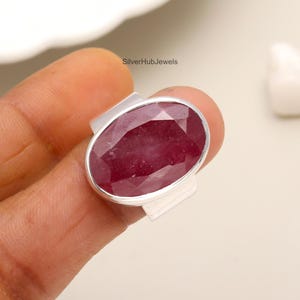 Könnte beinhalten: Ein silberner Ring mit einem großen, ovalen, roten Rubin-Edelstein. Der Ring hat ein schlichtes, modernes Design.