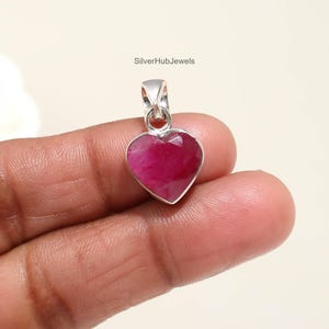 May include: A heart-shaped pendant with a faceted, deep pink gemstone set in a silver bezel. The pendant has a small loop for a chain. The text "SilverHubJewels" is visible.