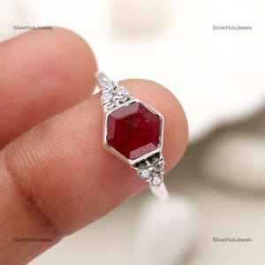 May include: A silver ring featuring a red hexagonal gemstone surrounded by smaller white gemstones.