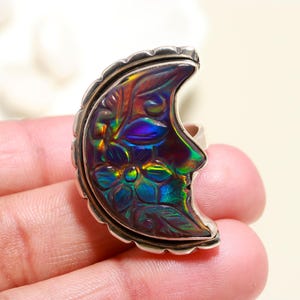 May include: A silver ring with a crescent moon design. The moon is carved with floral details and iridescent colours of blue, green, and purple. The ring has a textured border and a polished finish.