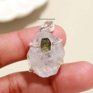 May include: A clear quartz pendant with a silver bail and wire setting. The pendant features a rough-cut, olive-green gemstone at its centre. The jewelry is designed to be worn as a necklace.