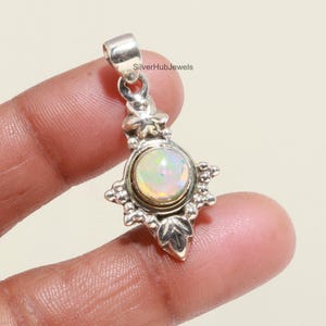 May include: A silver pendant with an oval opal gemstone. The pendant has a decorative floral design around the opal and a loop at the top for a chain. The text "SilverHubJewels" is visible on the top of the pendant.