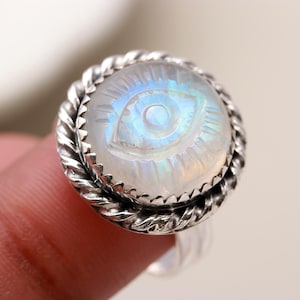 May include: A silver ring featuring a white moonstone eye design. The ring has a rope-like band.