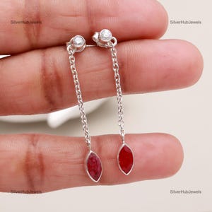 May include: A pair of silver drop earrings. Each earring features a small, clear gemstone at the top, connected to a chain that leads to a red, marquise-shaped gemstone. The earrings are displayed against a neutral background.