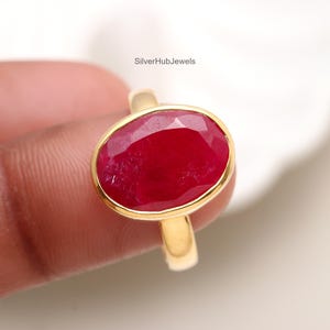 May include: A gold ring featuring a large, oval-cut ruby gemstone. The ruby is a deep red color and is set in a gold bezel. The ring band is also gold. The text "SilverHubJewels" is visible.