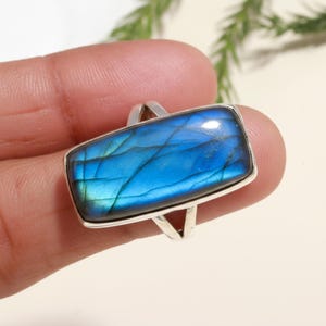 May include: A rectangular ring with a vibrant blue gemstone. The stone has a shimmering, iridescent quality with streaks of blue and green. The ring's band is silver-colored and features a split design.