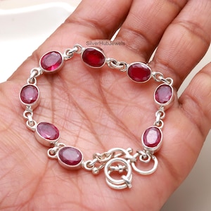 Könnte beinhalten: Ein silbernes Armband mit einem Schiebeverschluss verfügt über ovale, rote Edelsteine. Das Armband befindet sich an einem Handgelenk.