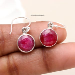 May include: A pair of sterling silver earrings with red ruby gemstones. The earrings have a simple drop design with a round ruby gemstone hanging from a silver hook.