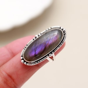 May include: A silver ring featuring an oval-shaped labradorite gemstone. The stone displays a mix of purple and gray hues. The ring has a decorative border and a simple band.