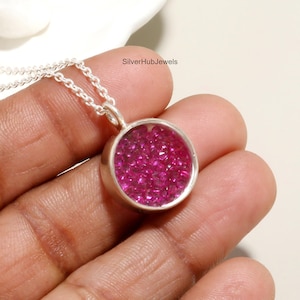 May include: A silver necklace with a round pendant filled with small, vibrant pink gemstones. The pendant is set in a silver bezel and hangs from a delicate silver chain. The text "SilverHubJewels" is visible.