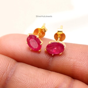 May include: A pair of gold stud earrings, each with a vibrant oval-cut ruby gemstone. The earrings are set in a classic prong setting, highlighting the rich red colour of the rubies. The text "SilverHubJewels" is visible.