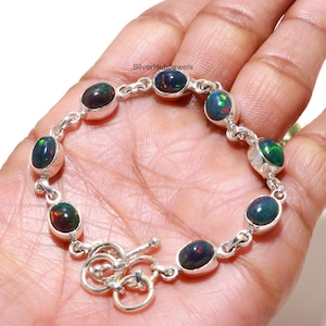 May include: A silver bracelet featuring seven oval-shaped dark black opals with iridescent flashes of green and red. The stones are set in smooth silver bezels and connected by silver links, finished with a circular toggle clasp closure.